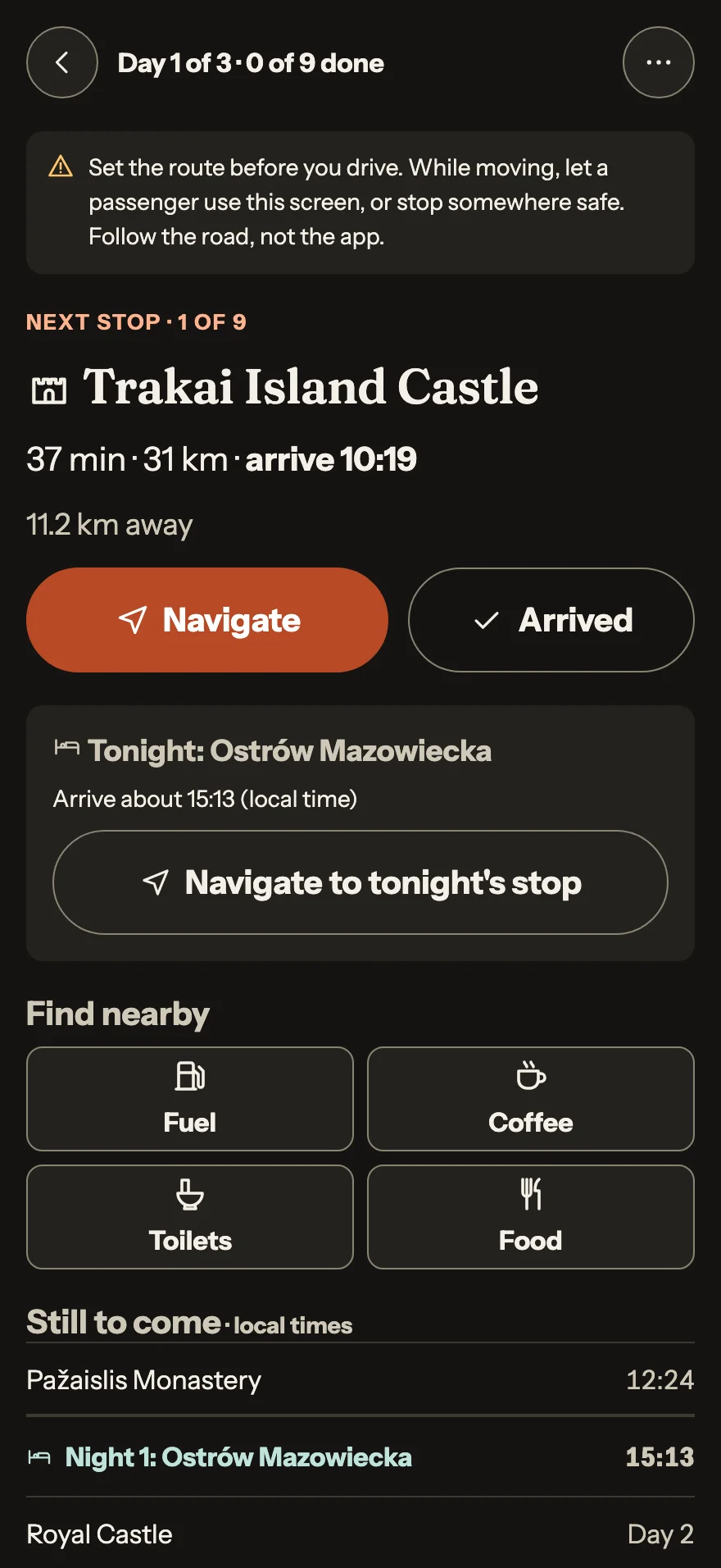 Dark drive-mode screen: Day 1 of 3, next stop Trakai Island Castle arriving at 10:19, tonight in Ostrów Mazowiecka, with buttons to find fuel, coffee, toilets and food.